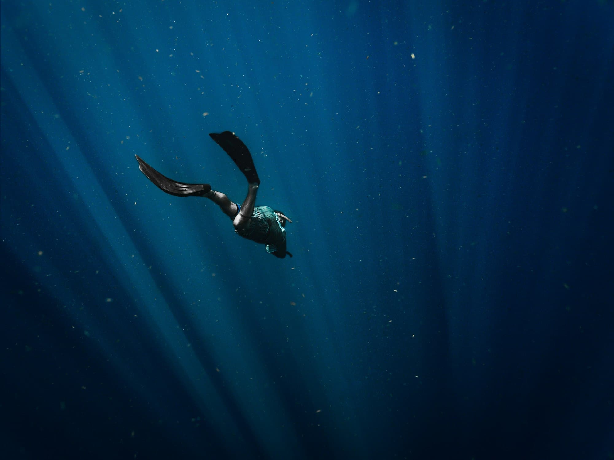 A freediver finning down into deep blue water, sunlight raking the surface above